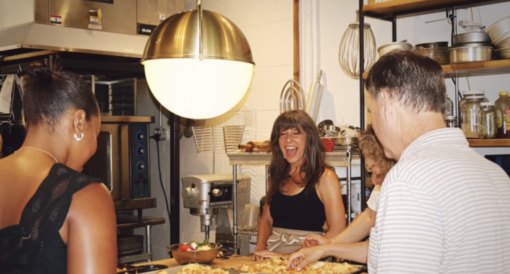 A diverse group of people stand around a table, smiling and laughing over a spread of food 